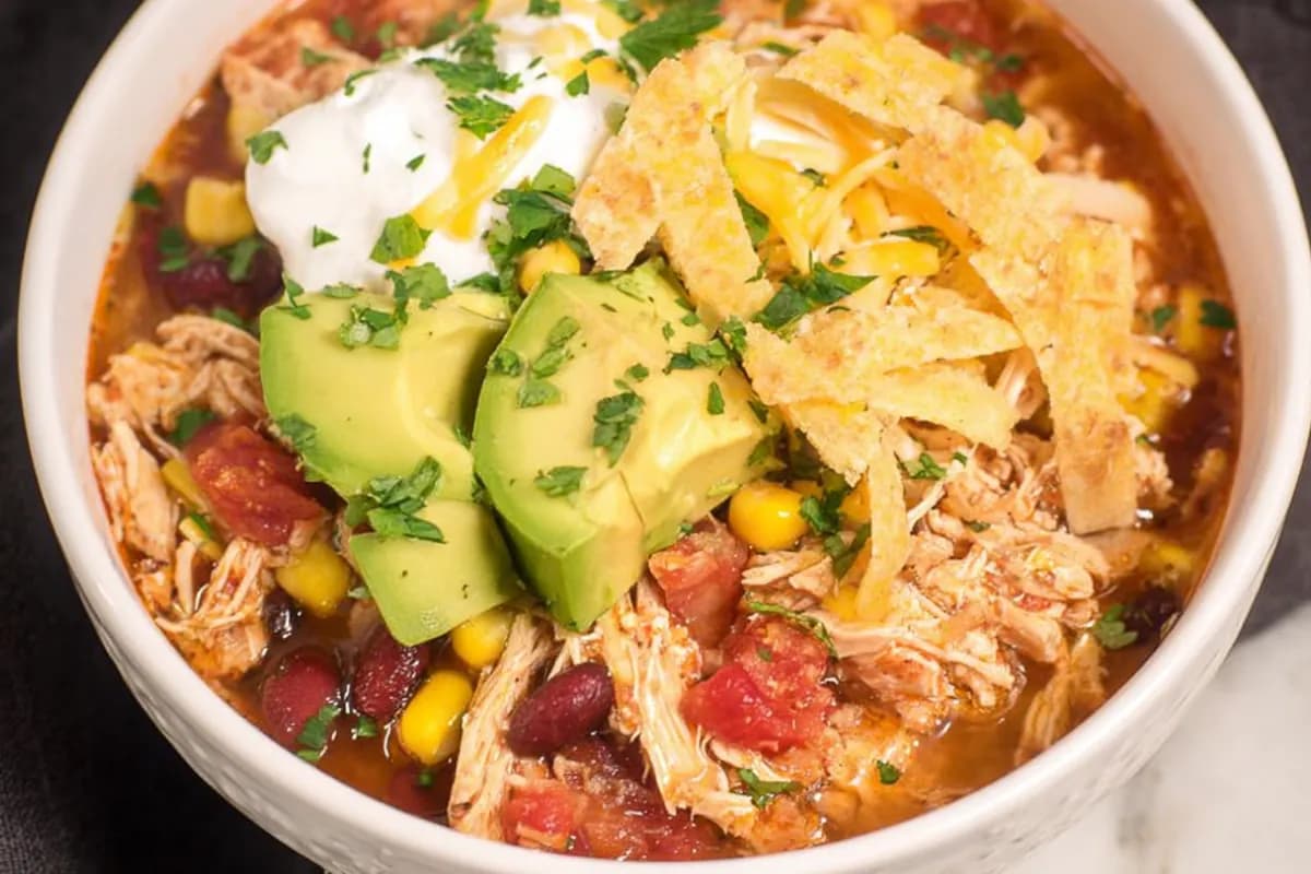 Chicken taco soup in a white bowl topped with avocado, sour cream, shredded cheese, and tortilla strips