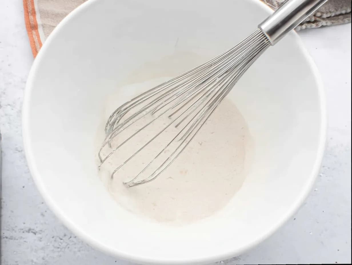Whisk mixing buttermilk, vanilla, and oil in a white bowl on a marble surface.